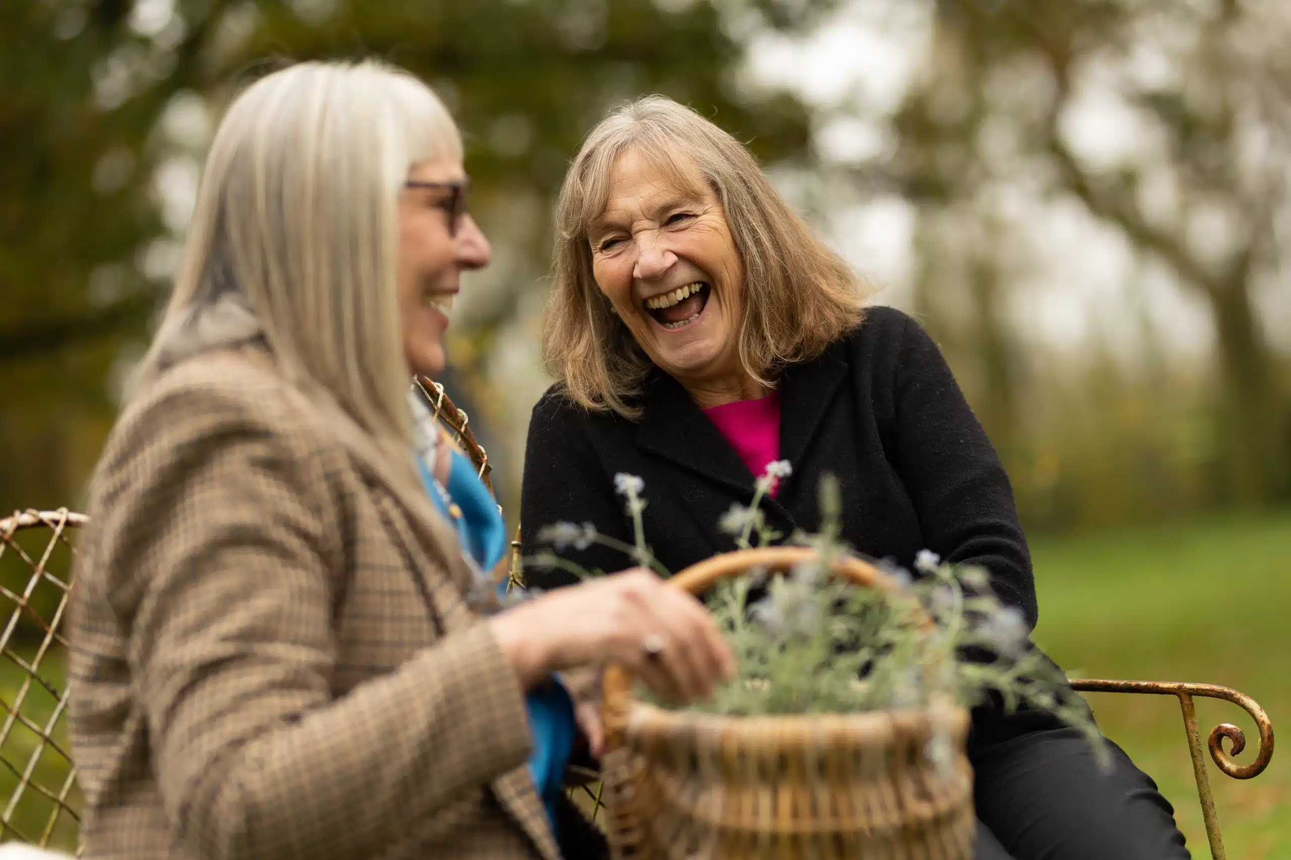 Two older women sit outdoors on a bench, smiling and laughing together. One holds a basket with flowers. Trees and greenery are visible in the blurred background, suggesting a park or garden setting.