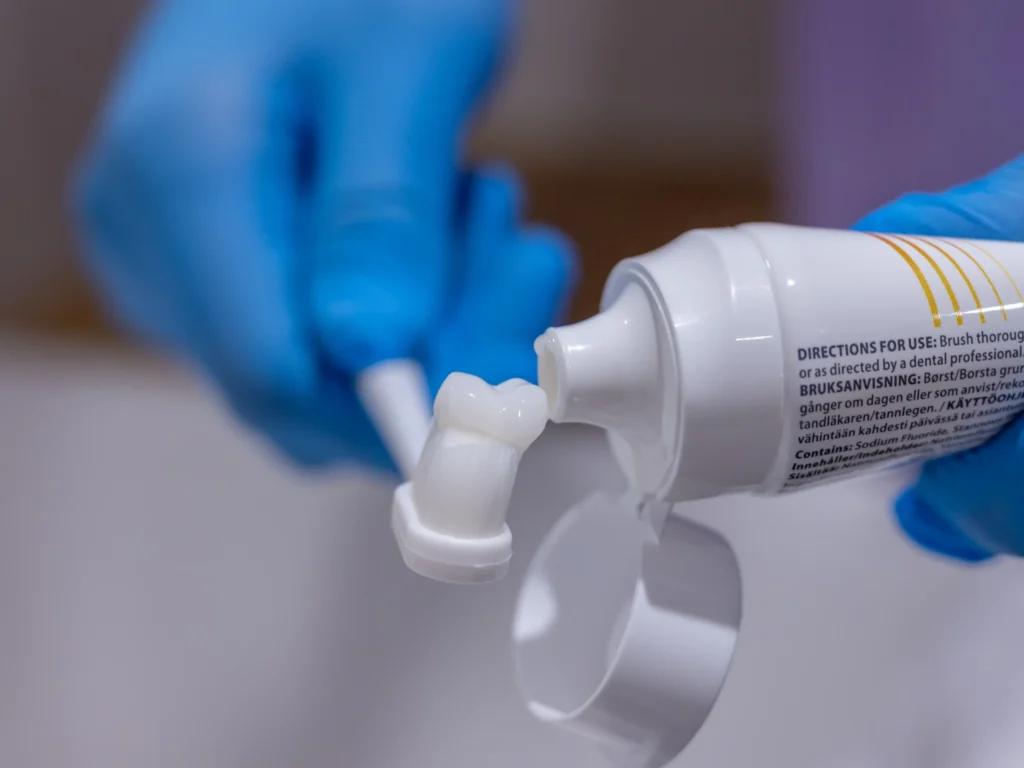 A person wearing blue gloves squeezes white toothpaste from a tube onto a toothbrush. The tube’s label with usage directions is partially visible. The toothbrush is held close to the toothpaste tube.