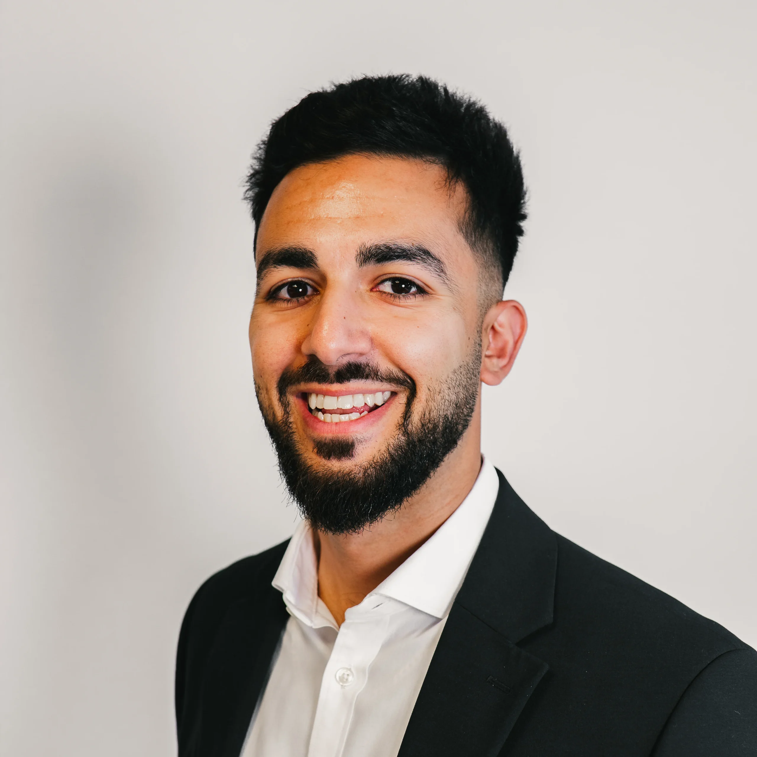 A man with short black hair and a trimmed beard is smiling, wearing a black suit jacket and a white shirt, standing against a plain light background.