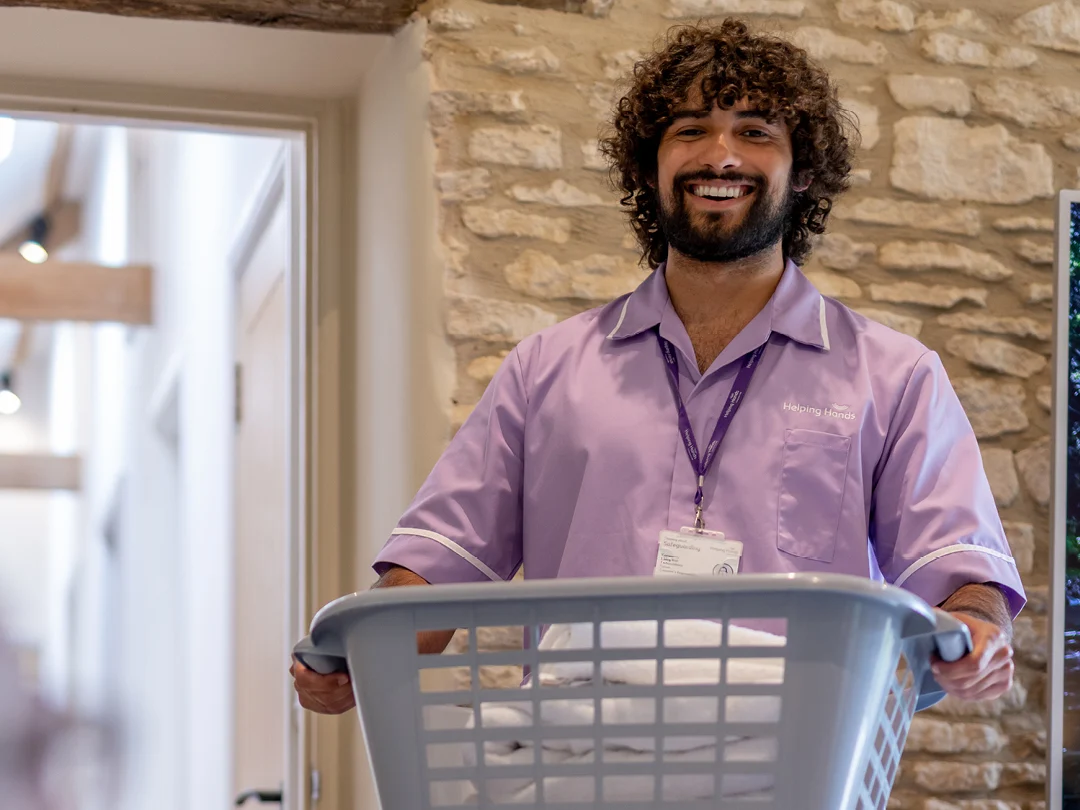 A smiling man in a lavender uniform holds a laundry basket filled with folded towels. He stands indoors near a stone wall, wearing an ID badge round his neck.