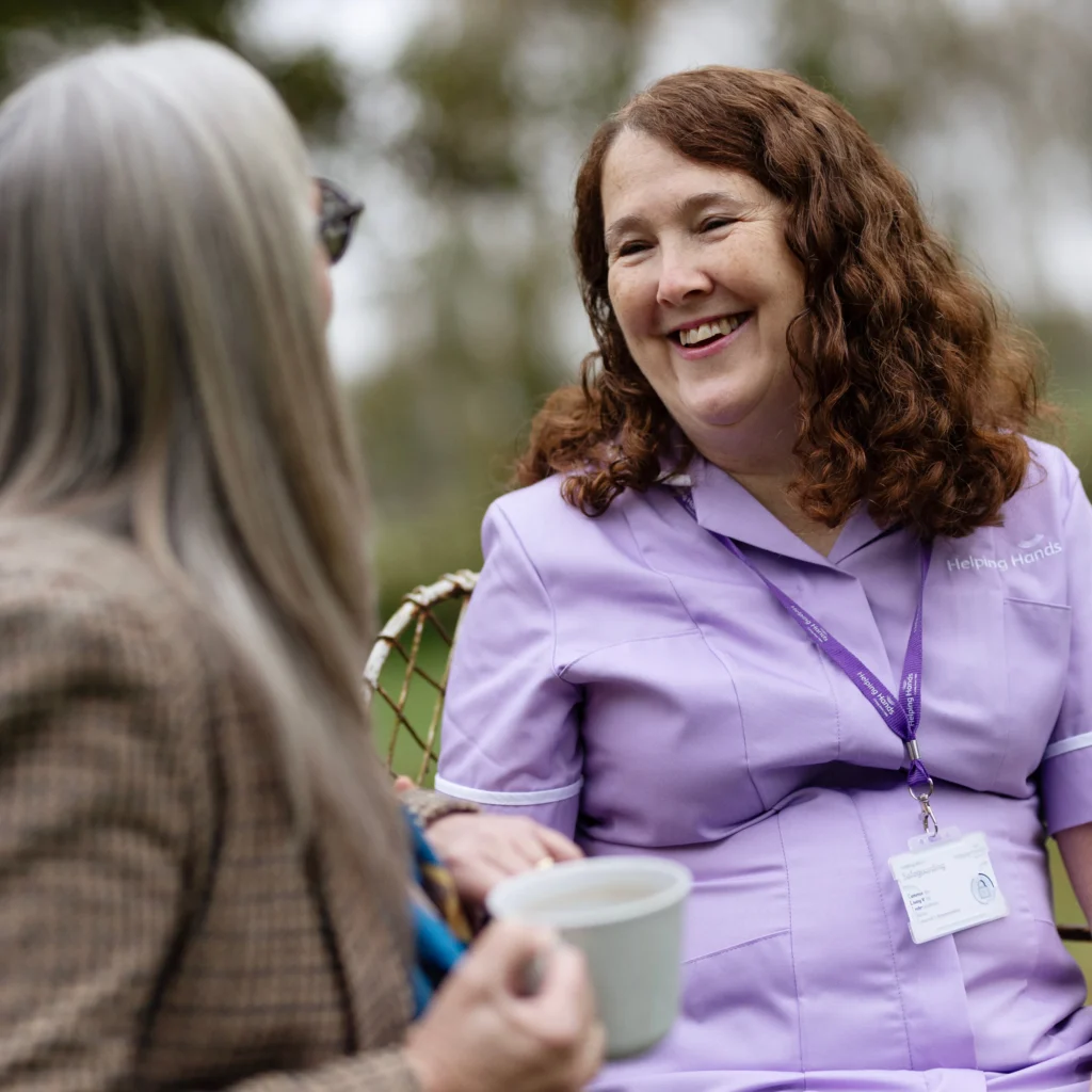 A smiling woman in a lavender uniform and name badge talks with an older woman holding a mug outdoors, suggesting a friendly conversation between a carer and her client.