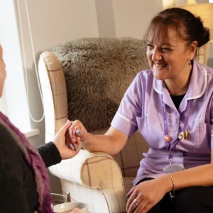 A smiling nurse in a purple uniform sits on an armchair, holding hands with another person, offering support and comfort in a cosy, warmly lit room.