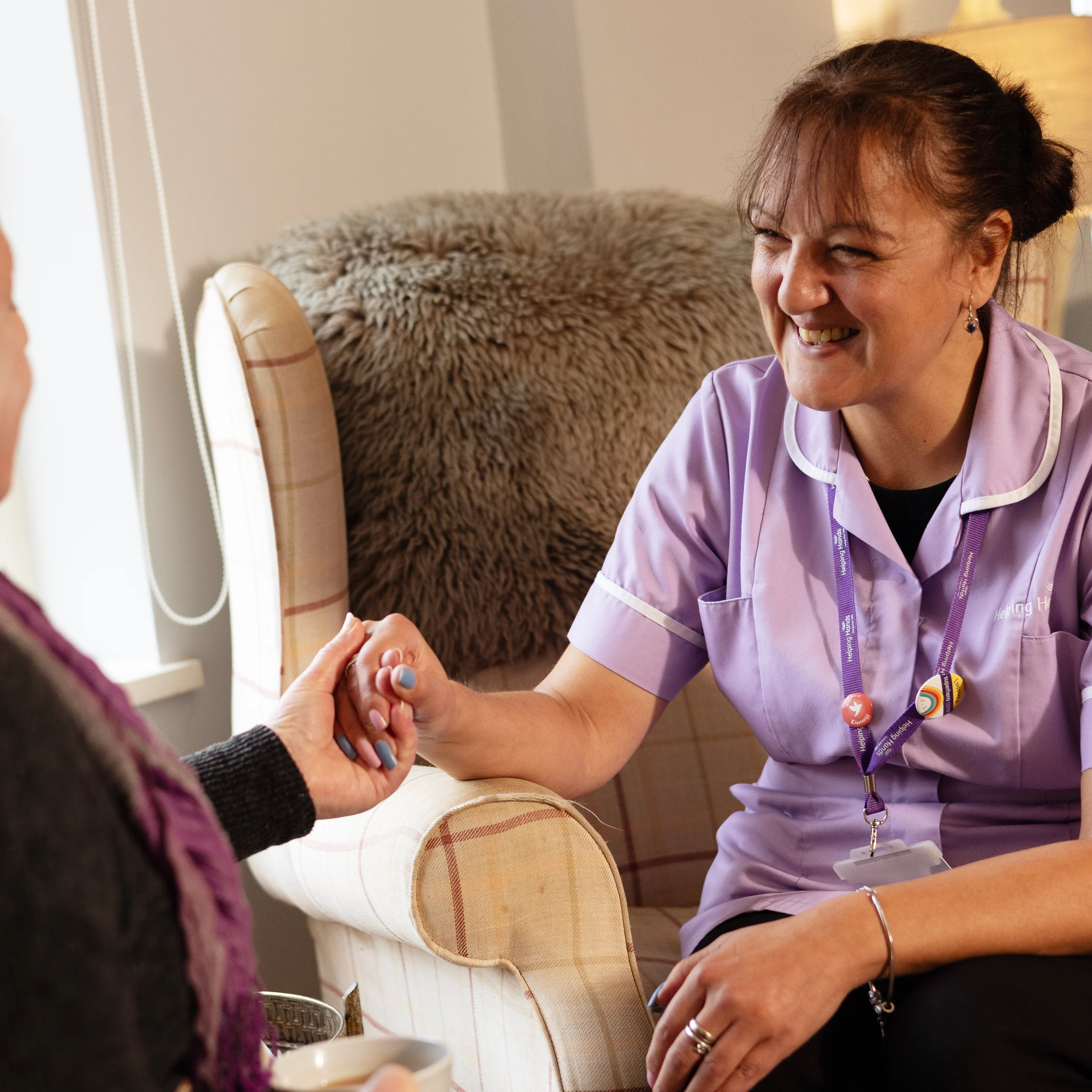A smiling nurse in a purple uniform sits on an armchair, holding hands with another person, offering support and comfort in a cosy, warmly lit room.
