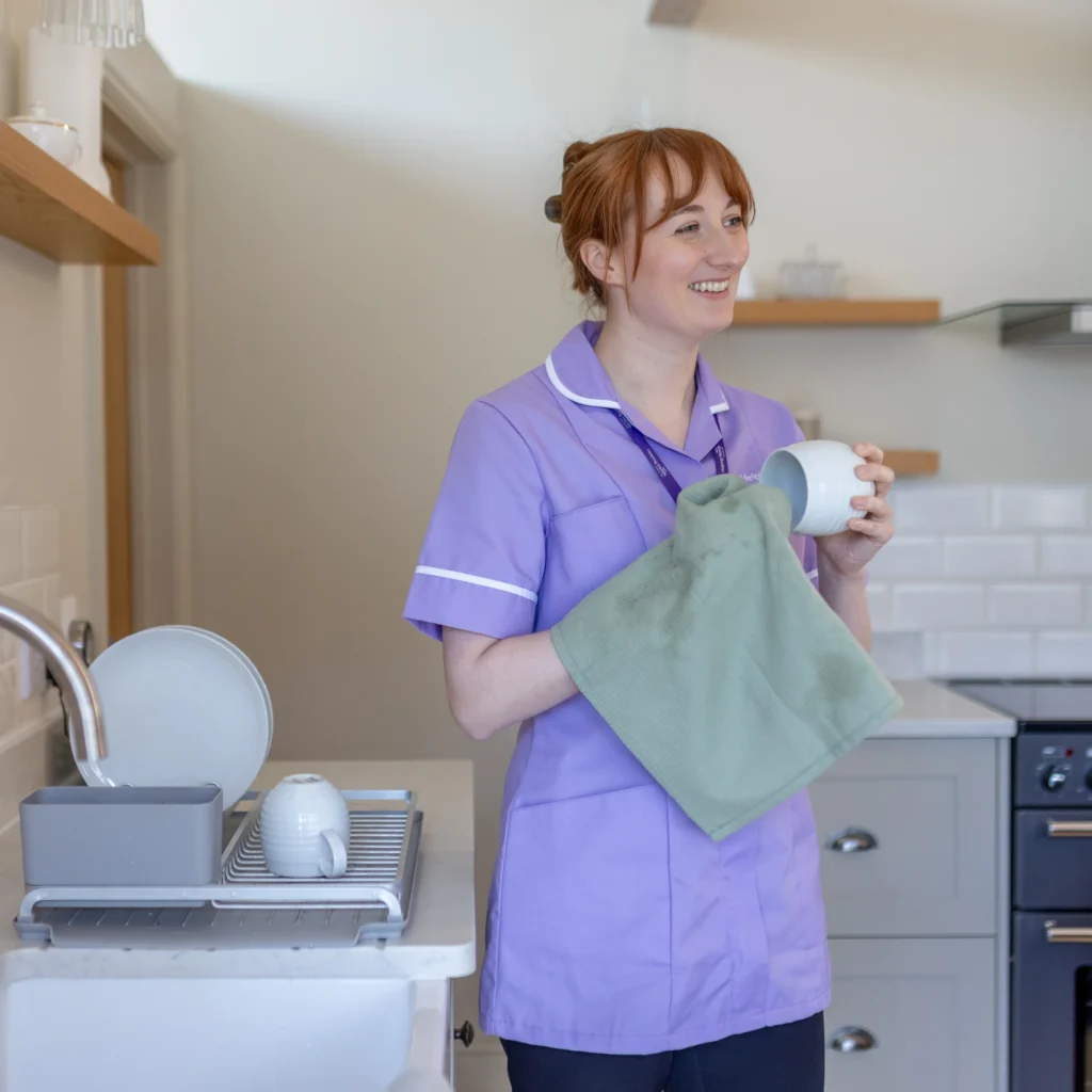 A smiling woman in a light purple uniform dries a white cup with a green tea towel in a modern kitchen. Clean dishes are stacked on a drying rack next to her. Shelves and cupboards are visible in the background.