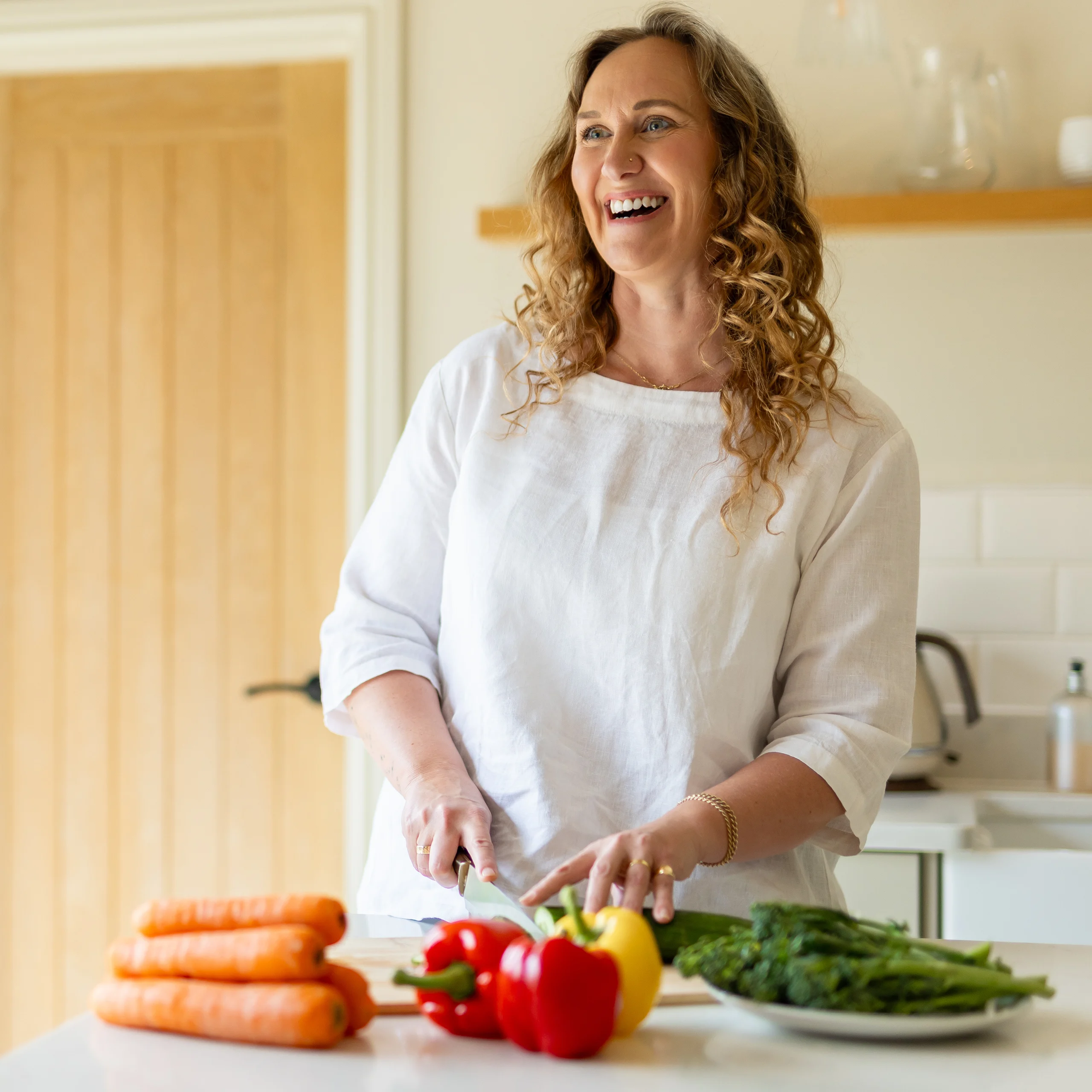A smiling woman with curly hair, wearing a white shirt, stands in a kitchen slicing vegetables. Carrots, peppers, and greens are arranged on the counter in front of her.