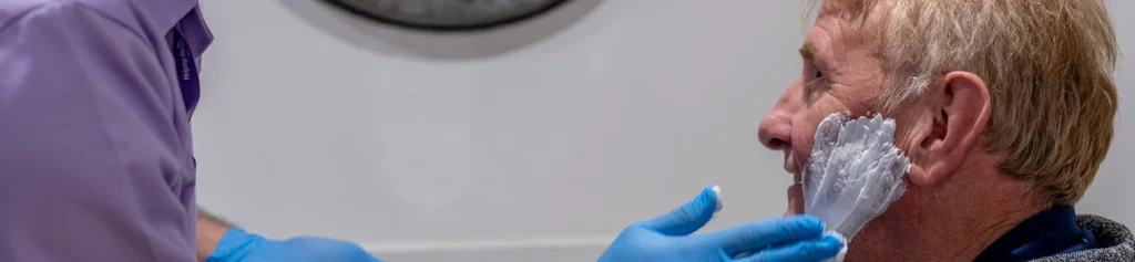 A person wearing blue gloves applies shaving foam to an older man’s cheek. The man is sitting and facing sideways, and part of a wall clock is visible in the background.
