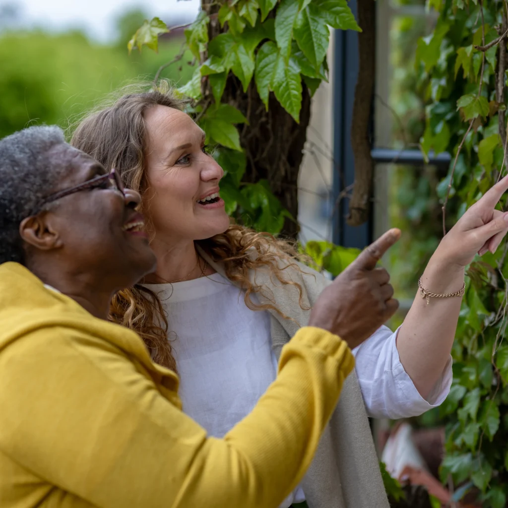 Two women smiling and pointing at green ivy on a garden wall, one wearing glasses and a yellow jacket, the other with long curly hair in a white shirt, enjoying time together outdoors.