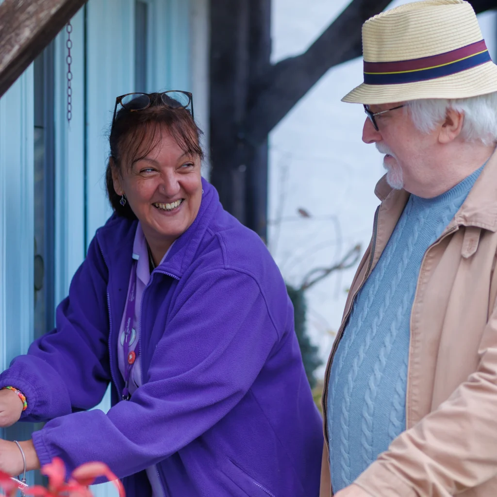 Two older adults smile and talk outside. The woman in a purple jacket stands near a door, while the man in glasses and a straw hat stands beside her, both appearing happy and engaged in conversation.