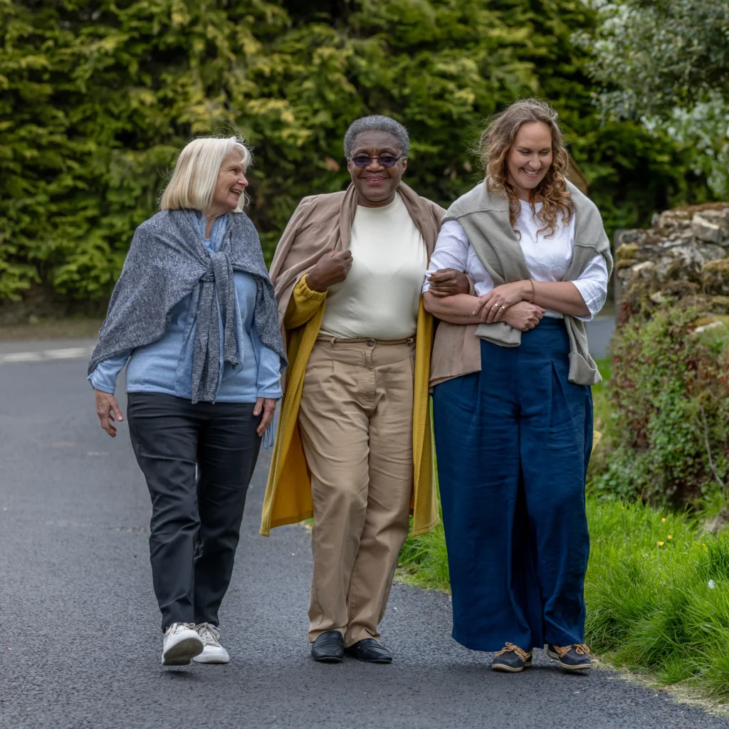 Three older women walk arm-in-arm down a paved path, smiling and enjoying each other's company, with greenery and trees in the background.