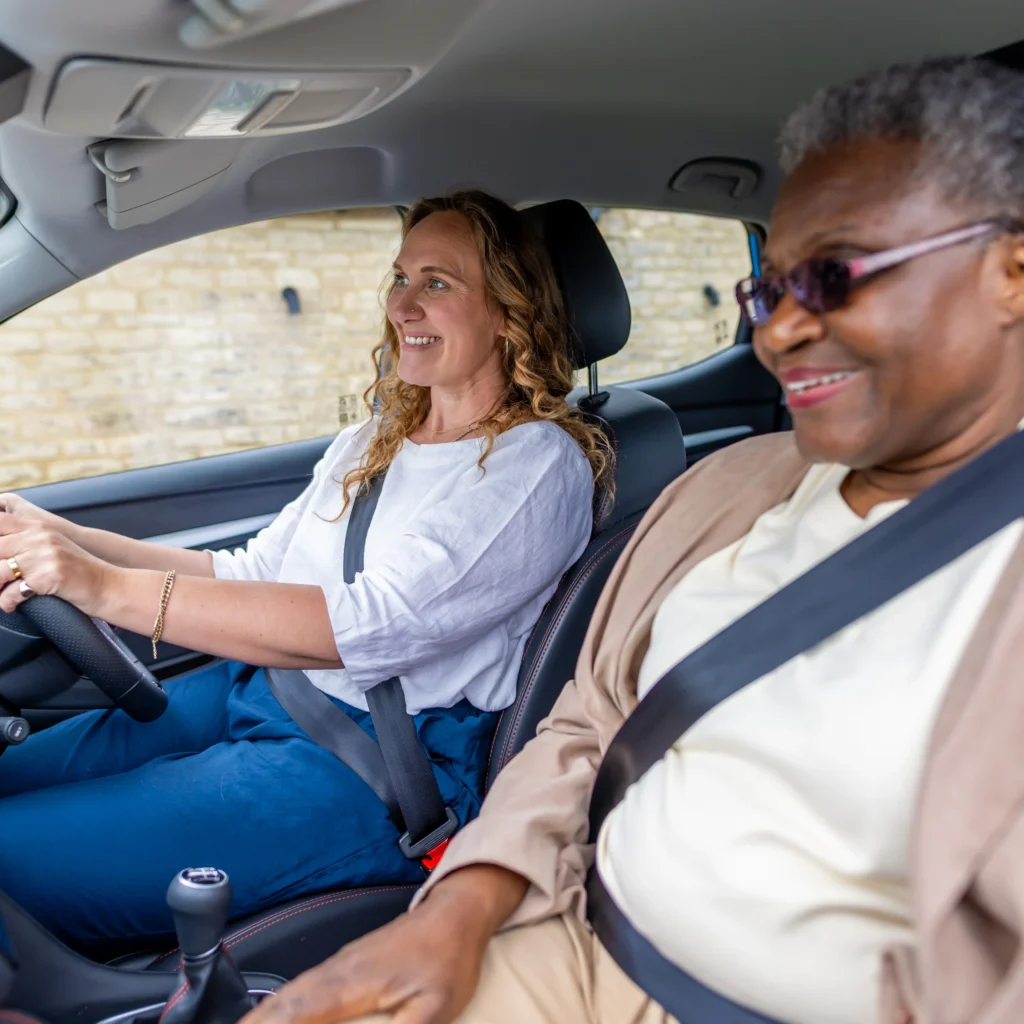 Two women sit in the front seats of a car, both wearing seatbelts. The woman driving has light skin and curly hair, whilst the passenger has dark skin, short grey hair, and sunglasses. Both appear to be smiling.