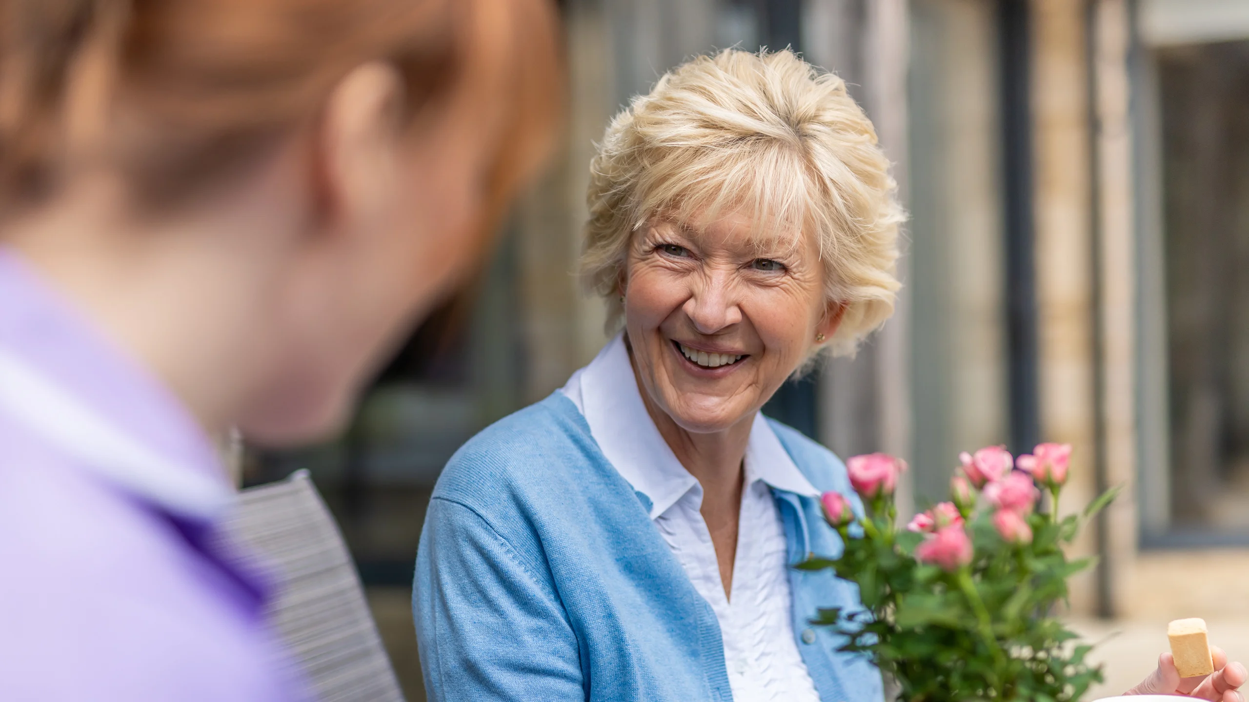 An older woman with short blonde hair smiles warmly whilst holding a bouquet of pink flowers, sitting outdoors and talking to another person whose face is blurred in the foreground.