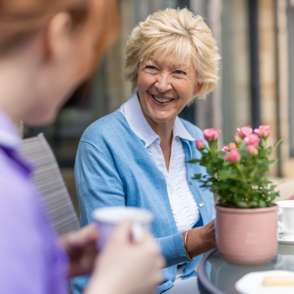 An older woman with blonde hair and a blue cardigan smiles whilst sitting at an outdoor table with a pink potted plant, talking to another person holding a mug.
