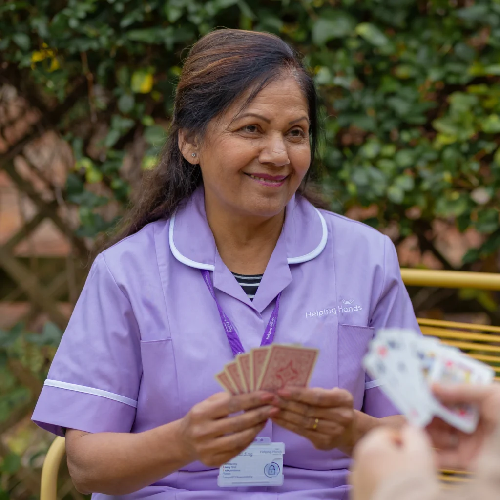 A smiling woman in a lavender uniform, holding playing cards, sits outdoors with another person. She has dark hair tied back and a name badge, and the background features greenery and a wooden trellis.
