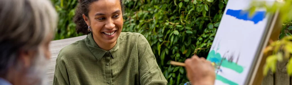 A woman smiles whilst sitting outdoors, watching someone paint on a canvas. Green foliage is visible in the background.
