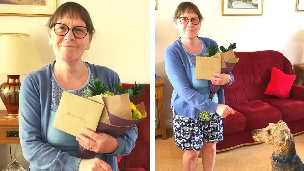 A woman with glasses and short hair holds flowers and a card, smiling inside a living room. In one image, she stands near a brown dog sitting in front of a red sofa.