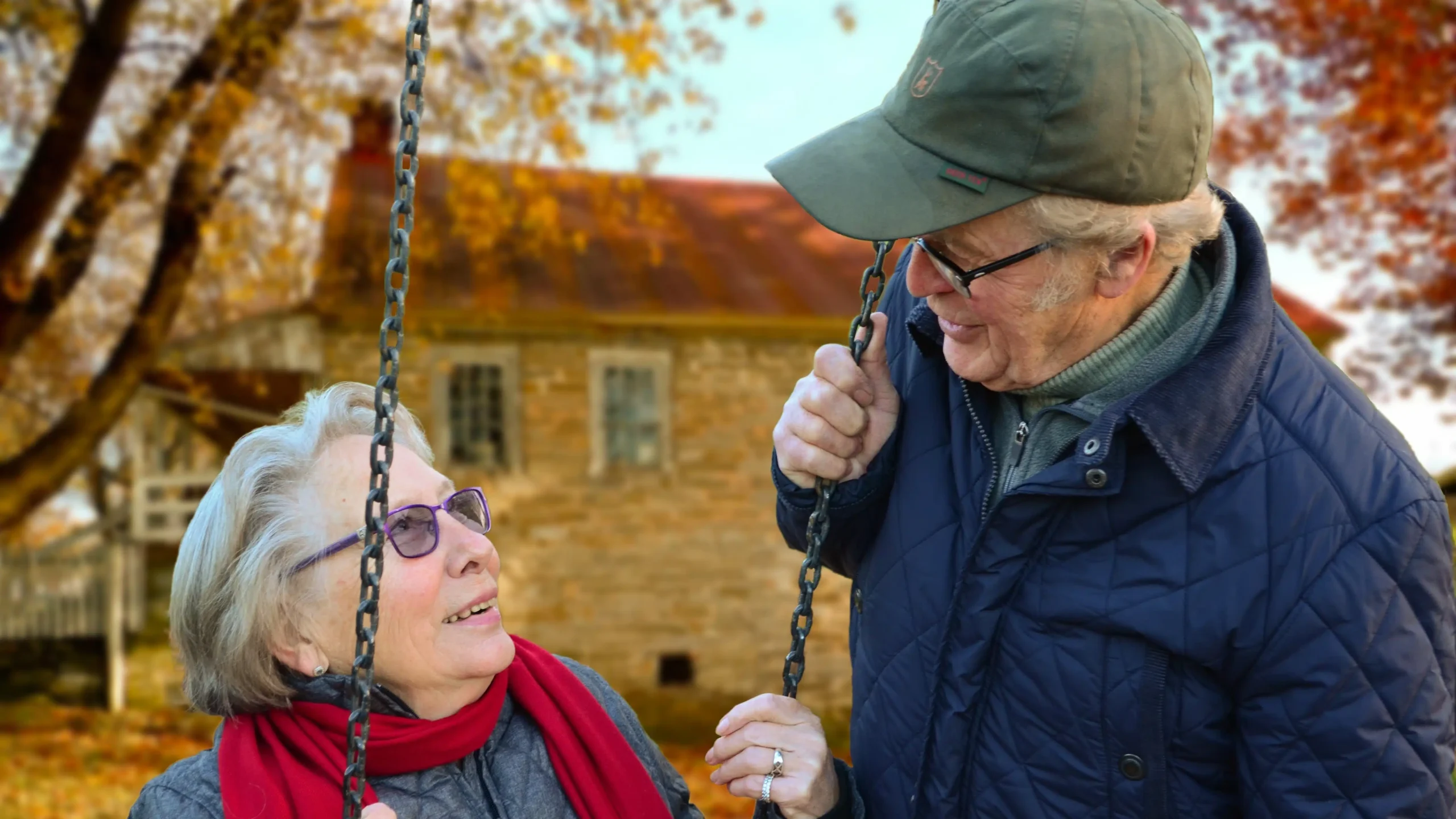 An elderly couple smile at each other while holding onto a swing outdoors, with autumn trees and a rustic house in the background. They are warmly dressed, wearing glasses and jackets.