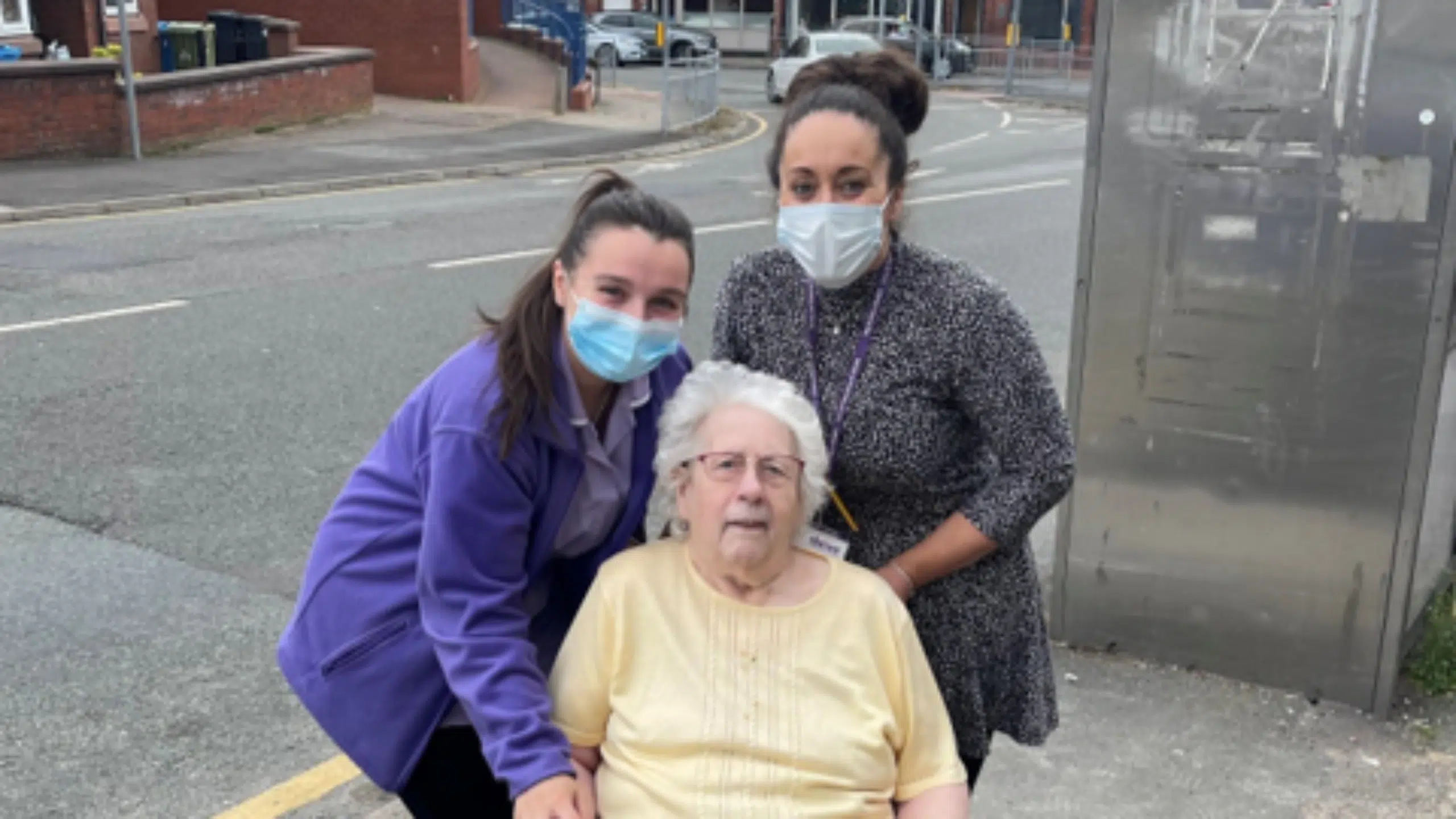 Two women wearing face masks stand outside on a street beside an elderly woman in a wheelchair, who is not wearing a mask. The group is smiling and posing for the photo on the pavement.