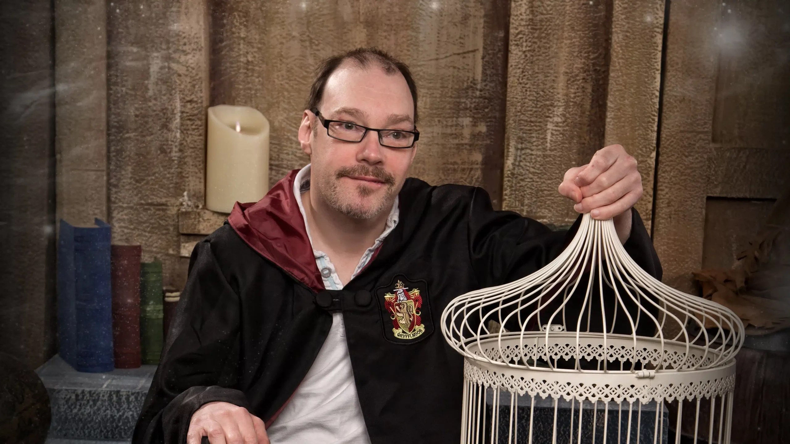 A man wearing glasses and a Gryffindor robe sits indoors, smiling and holding a decorative white birdcage. There is a large candle and stacked books in the background.