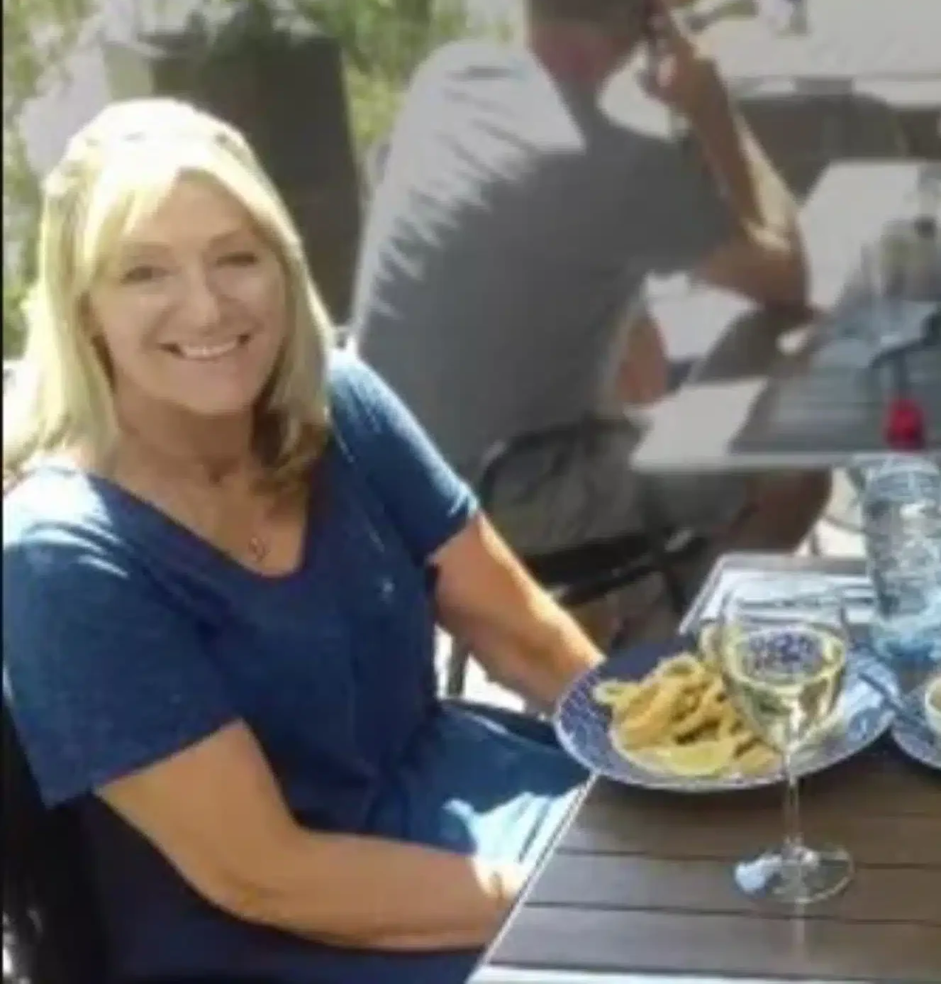 A woman with blonde hair in a blue shirt smiles at a table with a plate of food and a glass of white wine. In the background, a man sits at another table, talking on a phone.