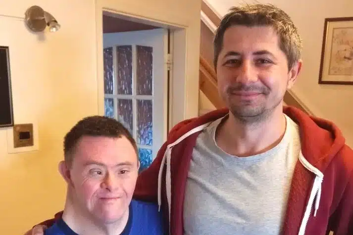 Two men stand indoors, smiling at the camera. The man on the left has Down’s syndrome and is wearing a blue shirt. The man on the right has short grey hair, a beard, and is wearing a red hoodie over a grey shirt.