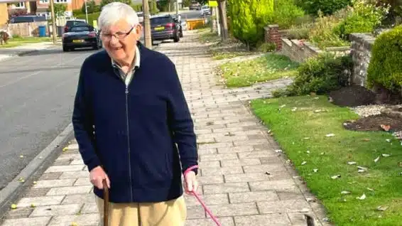An elderly man with white hair, glasses, and a navy jacket walks on a pavement using a walking stick and holding a small dog's pink lead on a suburban street.