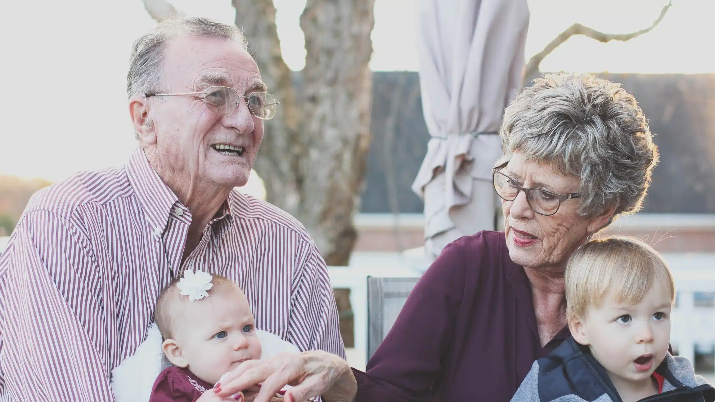 An elderly man and woman sit outdoors, smiling and holding two young children. The man, wearing glasses and a striped shirt, holds a baby; the woman, in a dark blouse, holds a toddler on her lap.
