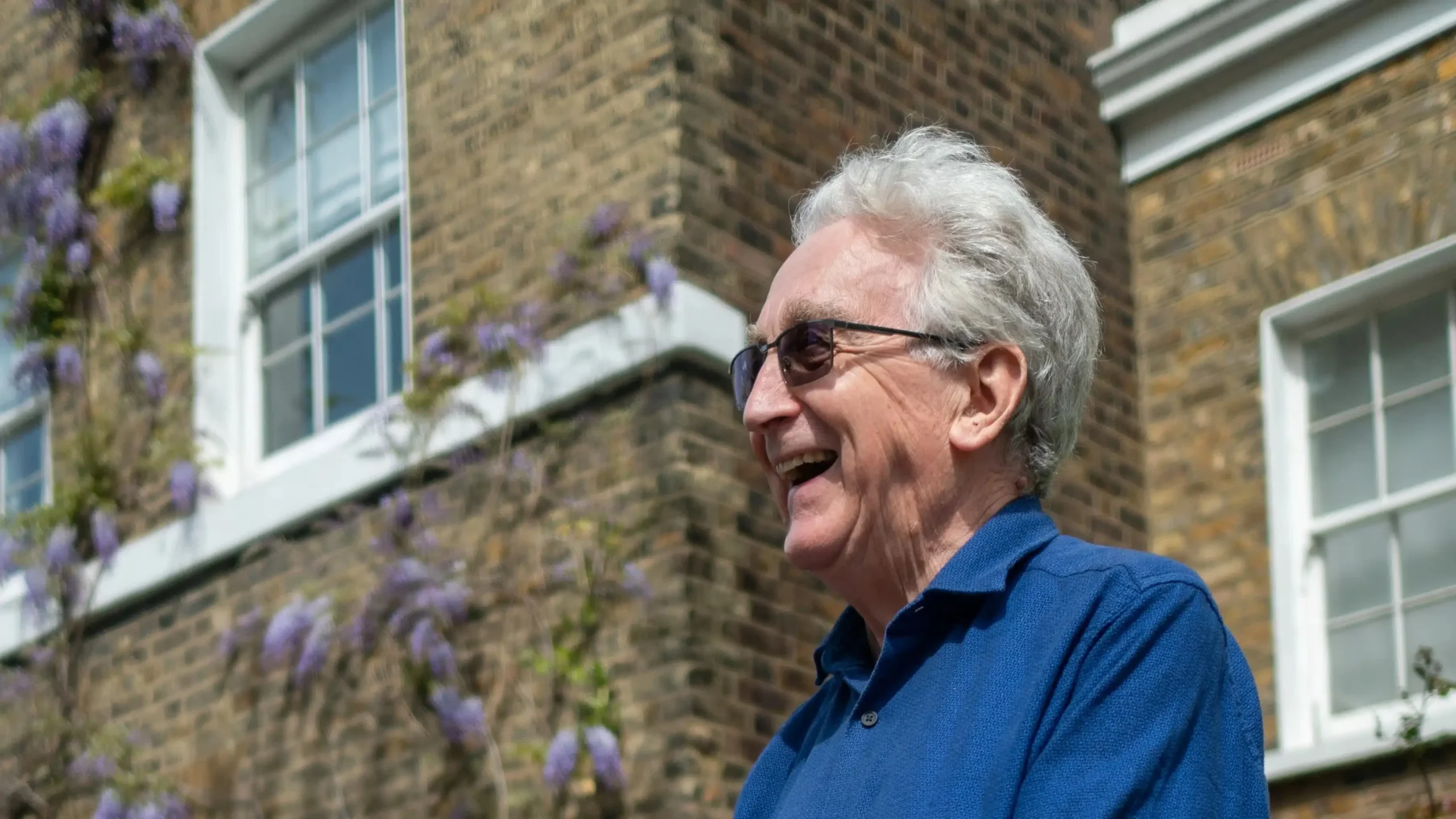 An older man with grey hair and glasses, wearing a blue shirt, stands outside a brick building, smiling and looking to the side. Purple flowers and a window are visible in the background.