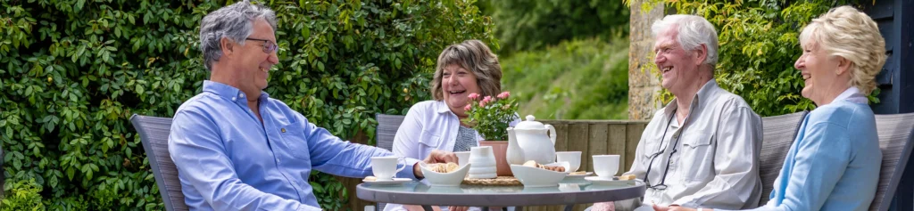Four older adults sit outside around a table, laughing and enjoying tea and snacks on a sunny day. The table has a teapot, cups, and plates of treats, with greenery and flowers in the background.