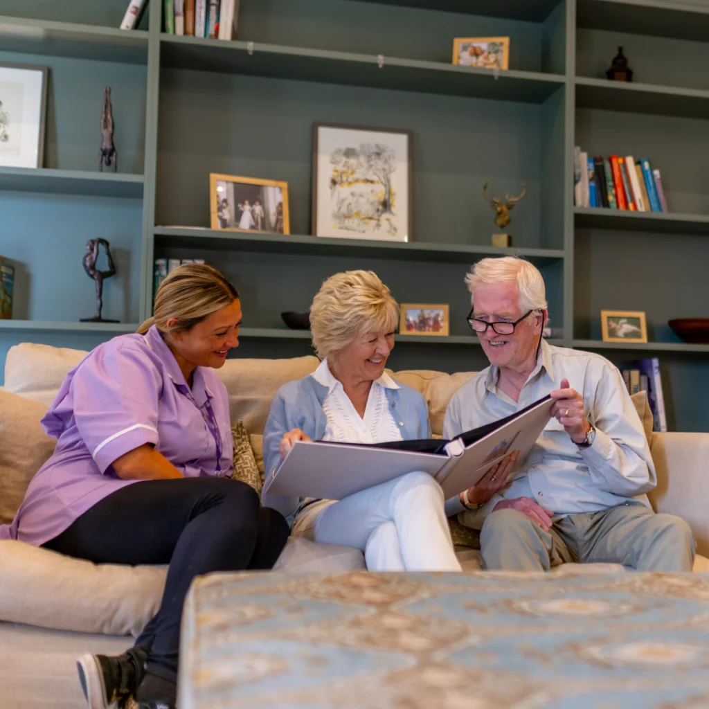 Three elderly people sit on a sofa, smiling and looking at a large photo album together, in a cosy living room with shelves and framed pictures in the background.
