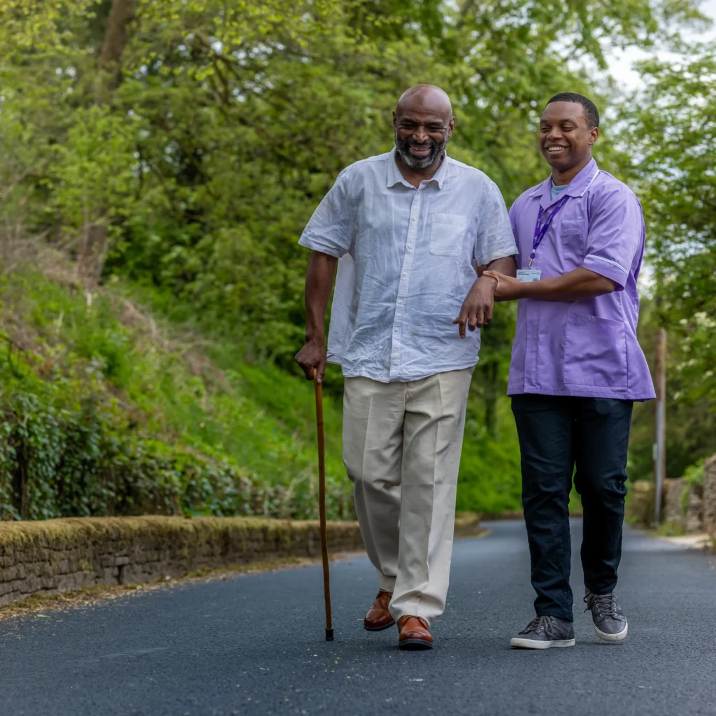 An older man using a walking stick walks outdoors on a paved path, smiling, while a younger carer in a purple uniform supports him. Trees and greenery line the path.