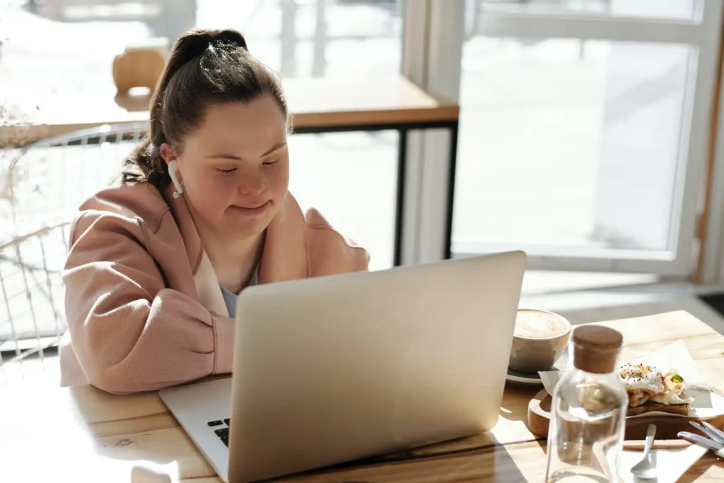 A young woman with Down’s syndrome sits at a table in a bright café, looking at a laptop. There is a cup of coffee and a plate of food beside her. Sunlight streams in through large windows in the background.