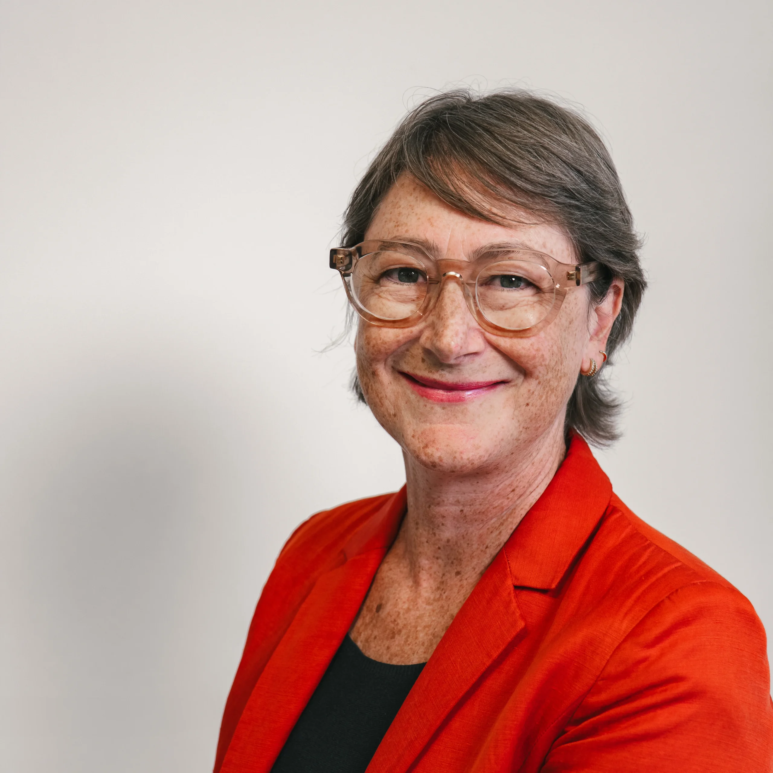 Smiling middle-aged woman with short brown hair, wearing clear glasses, a bright orange blazer, and a black top, standing against a plain light background.
