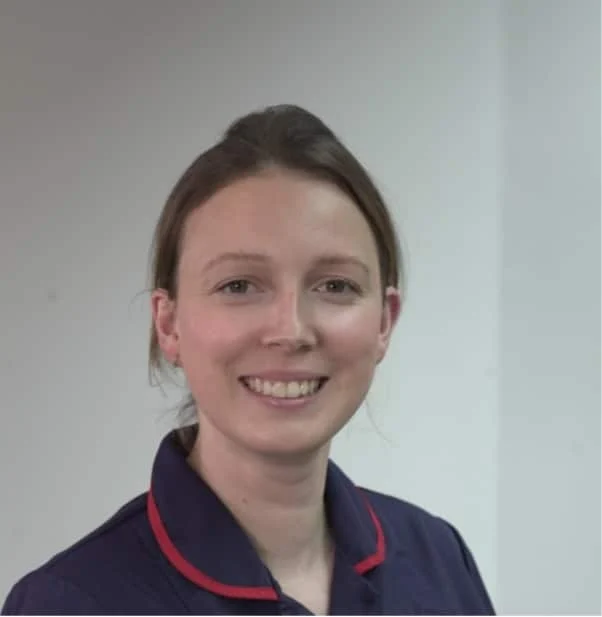 A woman with light brown hair, tied back, smiles at the camera. She is wearing a dark shirt with a red-trimmed collar and stands in front of a plain light-coloured background.