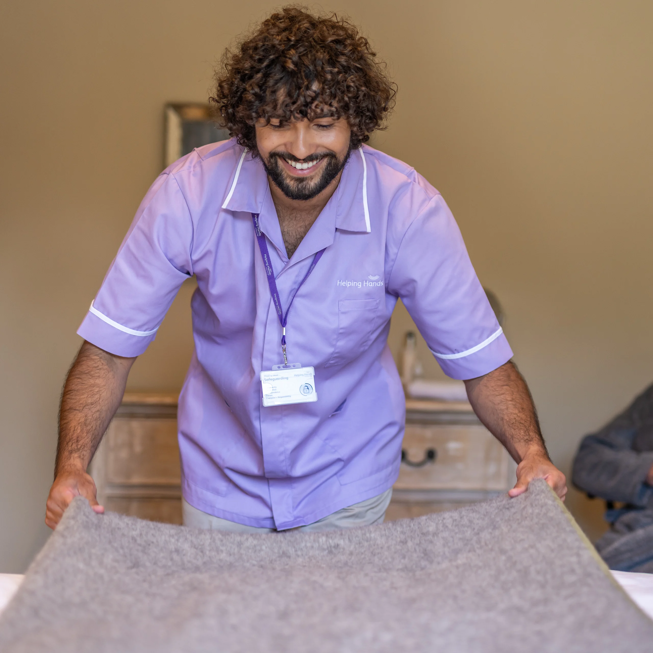 A smiling man with curly hair, wearing a light purple uniform and name badge, is making a bed, spreading a grey blanket over it in a warmly lit room.