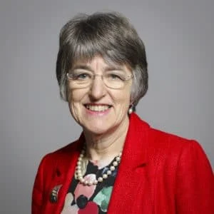 An older woman with short grey hair, glasses, and a friendly smile wears a bright red blazer over a colourful top, accessorised with a pearl necklace and brooch, posing against a plain grey background.