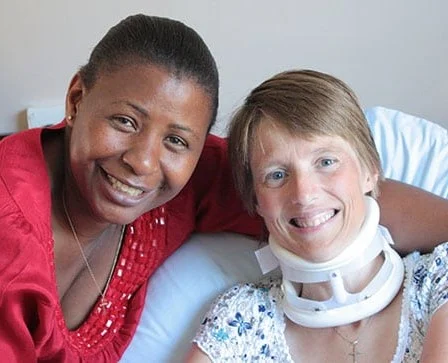 Two women smiling together; one wears a red top, and the other is lying on a bed with a white floral shirt and a medical neck brace. They appear happy and close, sharing a warm, supportive moment.