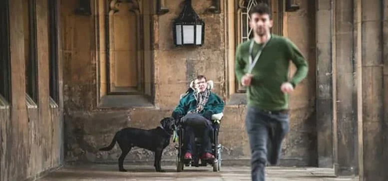 A person in a wheelchair, accompanied by a black guide dog, sits in a stone corridor. Another person in a green jumper is running towards the camera in the foreground.