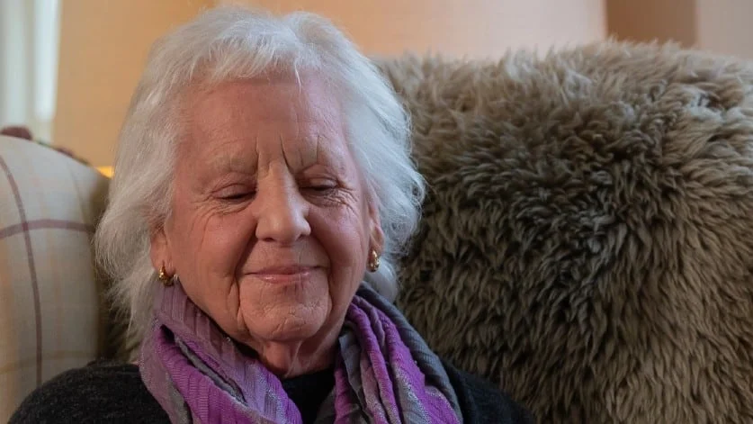 An older woman with white hair and gold earrings sits on a sofa, eyes closed, smiling gently. She is wearing a purple scarf and dark top, with a fluffy cushion behind her.