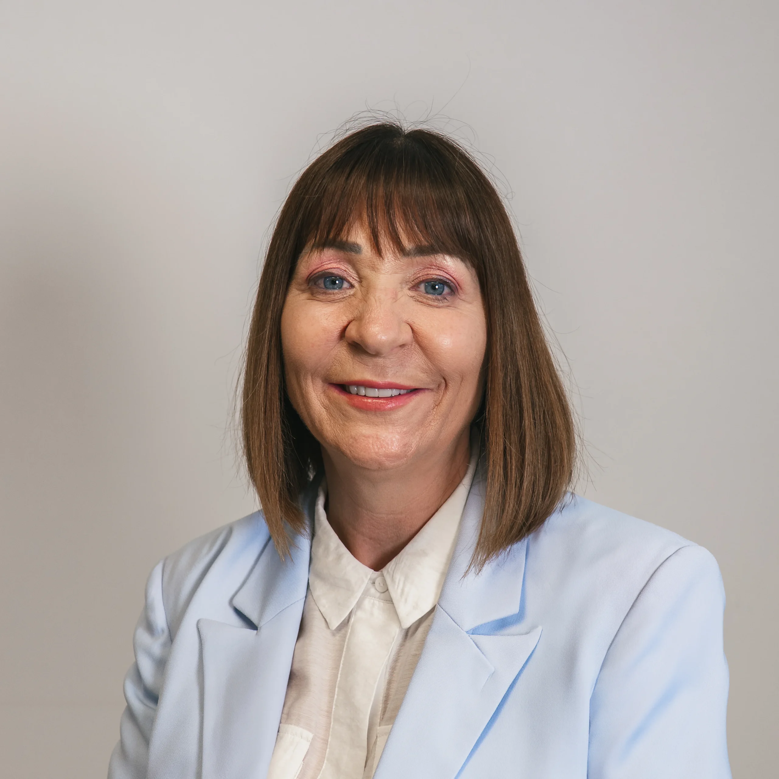 A woman with straight brown hair and a fringe is smiling at the camera. She is wearing a light blue blazer over a white collared shirt and is posed in front of a plain light grey background.