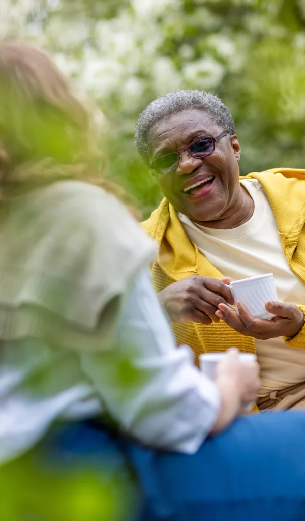 An older adult wearing glasses and a yellow jumper laughs whilst holding a cup, enjoying a conversation with another person outdoors. Lush greenery is visible in the background.
