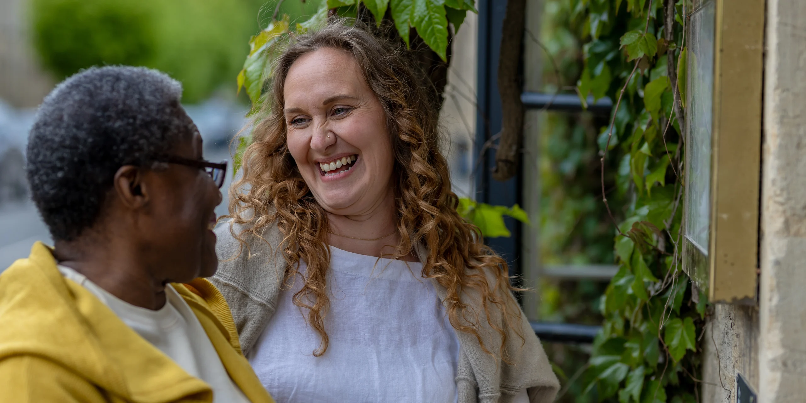 Two people stand outside by a wall covered in green leaves, smiling warmly at each other. One has curly hair and wears a light top, the other wears glasses and a yellow jacket. The atmosphere is cheerful and friendly.