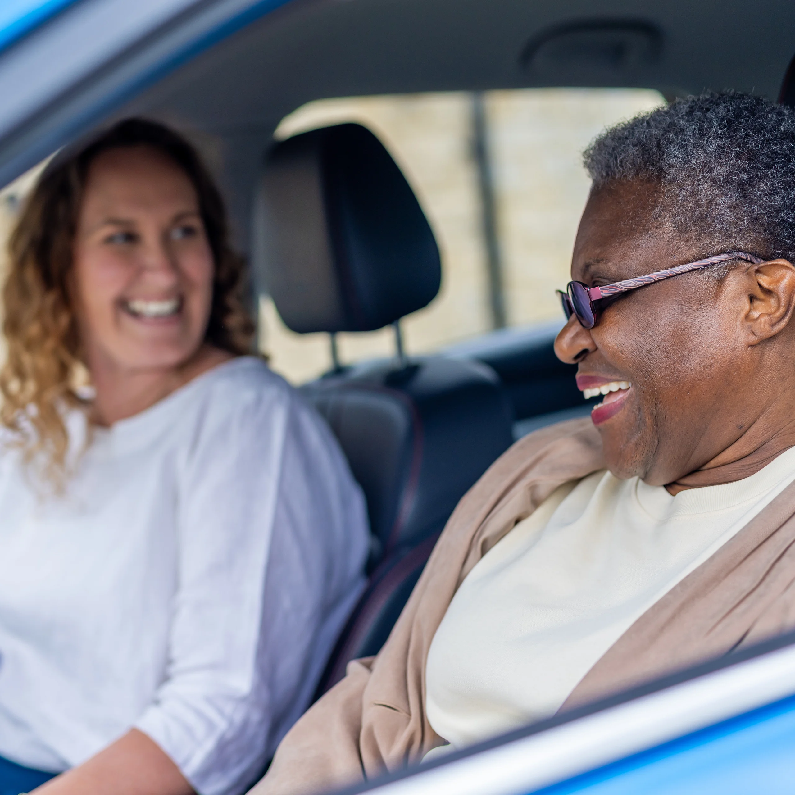 Two women sit in a car, smiling and laughing together. The woman in the driver's seat wears glasses and a beige jacket, whilst the passenger has curly hair and a white top. The mood is joyful and relaxed.