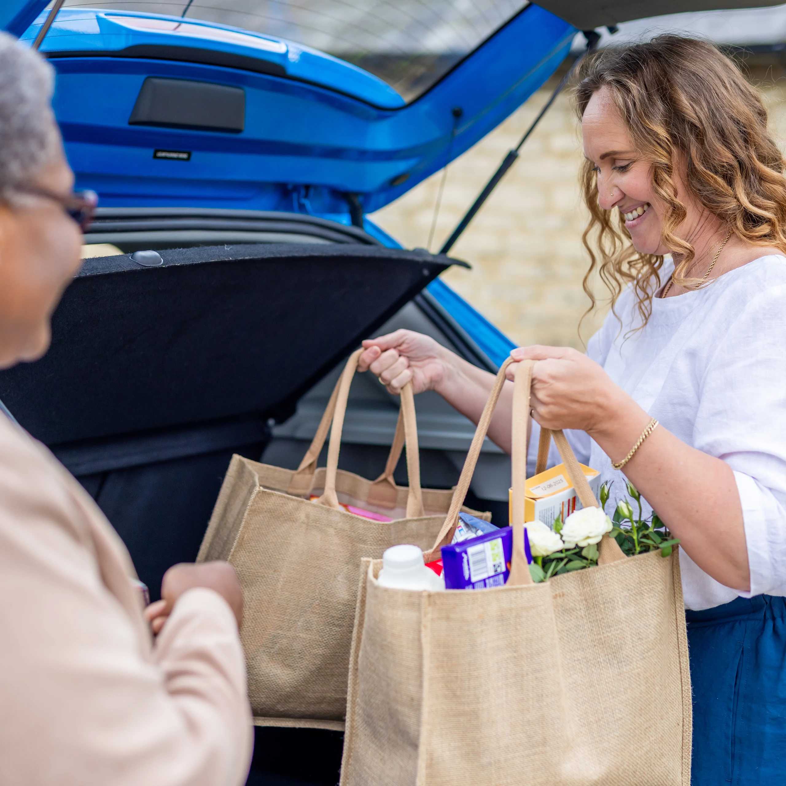 Two women stand by an open car boot, smiling as they hold reusable shopping bags filled with groceries, including flowers and a carton.
