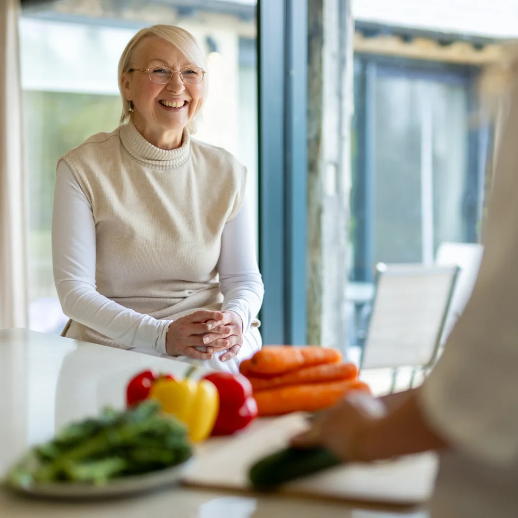 An older woman with glasses smiles while sitting at a kitchen counter with carrots, tomatoes, and a yellow pepper. Another person in the foreground is slicing a cucumber. Sunlight filters through large windows.