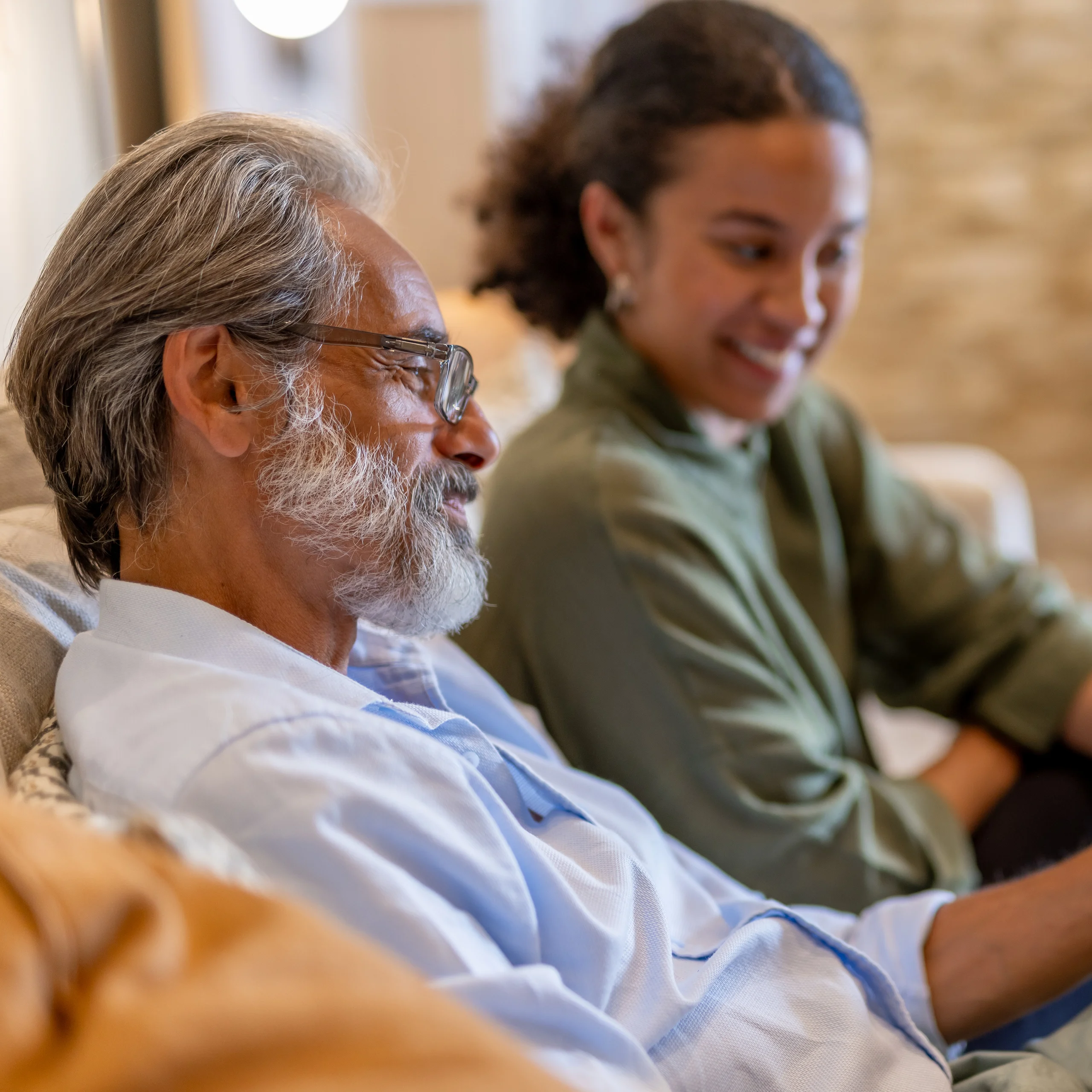 An older man with grey hair and glasses sits smiling on a sofa next to a young woman with curly hair, who is also smiling. They appear to be enjoying a pleasant conversation in a cosy indoor setting.