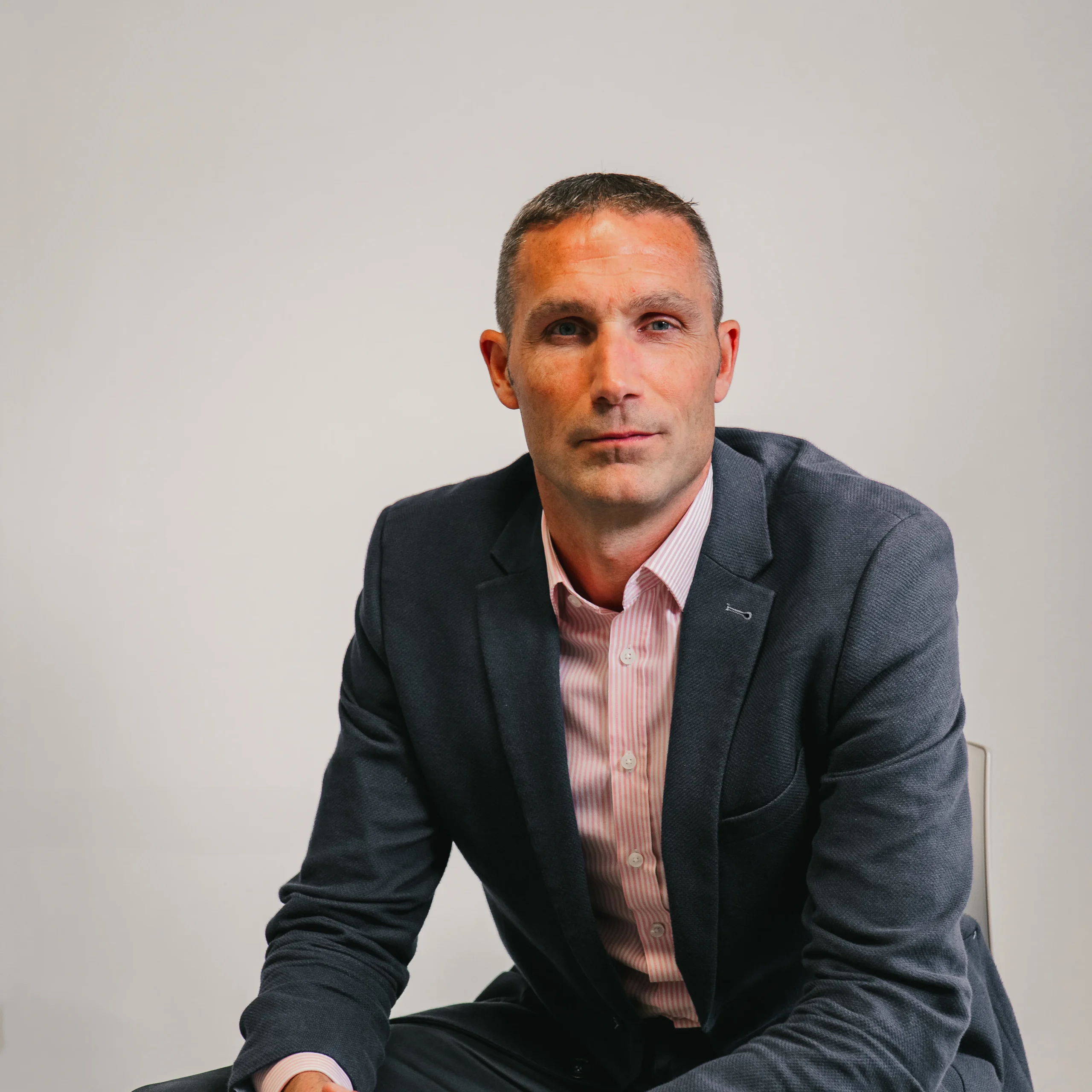 A man in a dark blazer and light pink shirt sits against a plain light background, looking directly at the camera with a neutral expression.