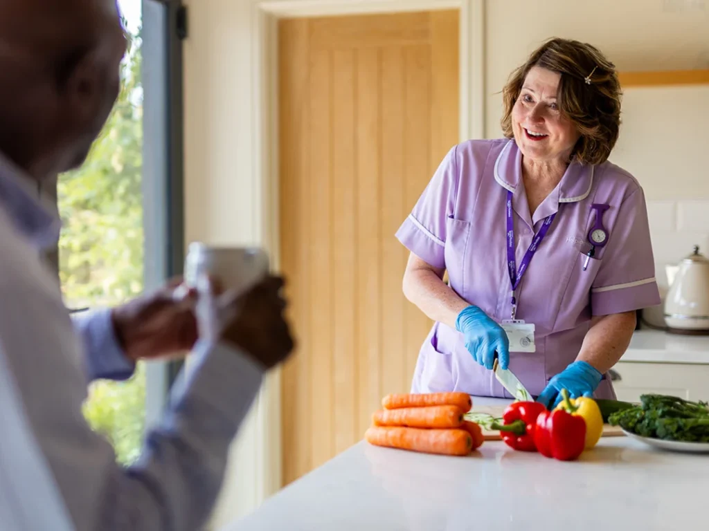 A nurse in a purple uniform and blue gloves smiles while chopping vegetables in a kitchen, talking to a person holding a mug in the foreground. Carrots and peppers are on the counter.