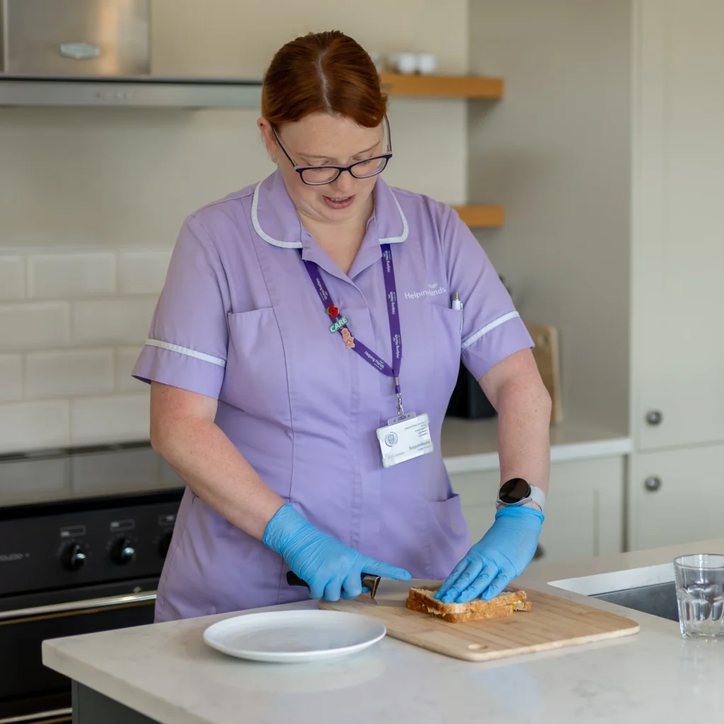 A woman in a light purple uniform and blue gloves slices bread on a wooden chopping board in a kitchen. She wears glasses and a name badge, and there is a plate and a glass of water on the worktop.