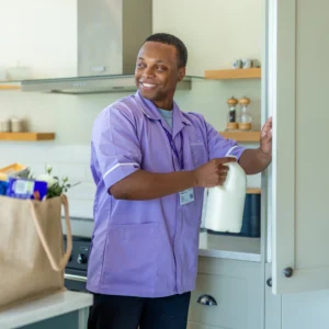 A smiling man in a light purple uniform places a milk bottle in a kitchen cupboard. A beige shopping bag with groceries sits on the worktop beside him. The kitchen has light cupboards and open shelves.