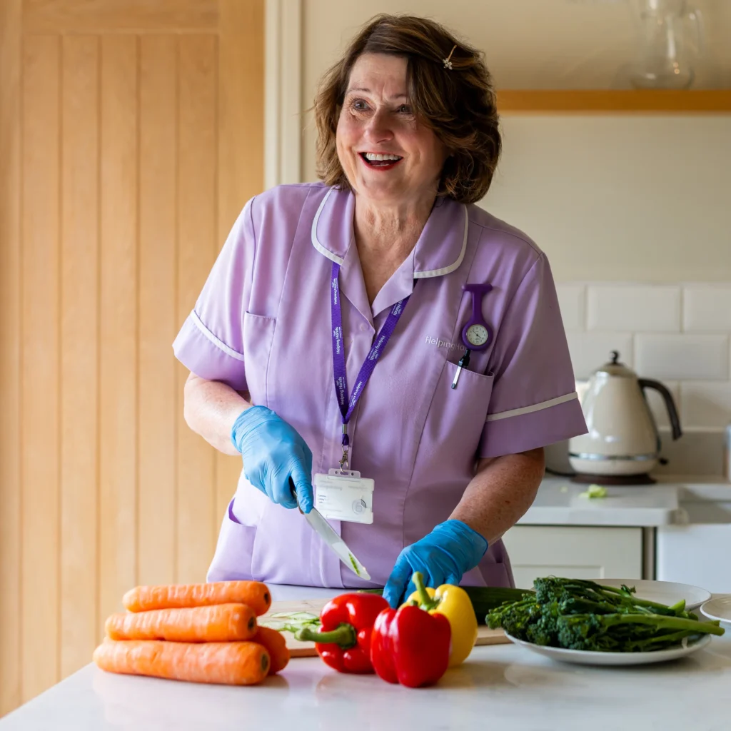 A woman wearing a lilac uniform and blue gloves smiles whilst chopping vegetables, including carrots and peppers, in a bright kitchen. A kettle and a plate of greens are visible on the worktop nearby.